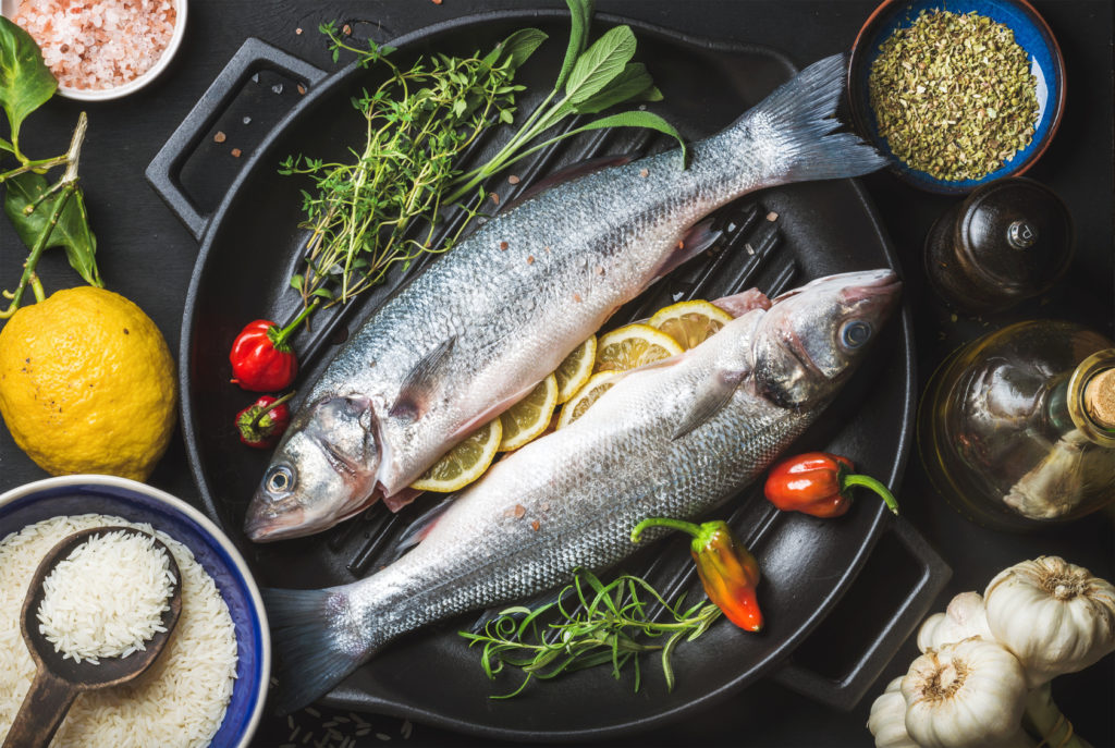 Ingredients for cooking healthy fish dinner. Raw uncooked seabass with rice, lemon, herbs and spices on black grilling iron pan over dark background