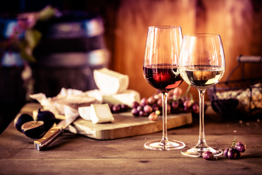Cheese platter with fresh grapes and glasses of red and white wine on a rustic wooden table in front of a blazing fire in a tavern or winery
