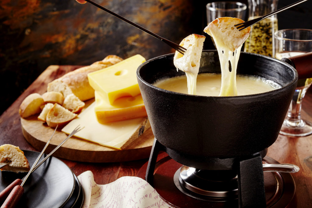 Gourmet Swiss fondue dinner on a winter evening with assorted cheeses on a board alongside a heated pot of cheese fondue with two forks dipping bread and white wine behind in a tavern or restaurant